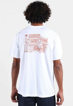 T-shirt en coton blanc avec une impression graphique douce au dos représentant une scène de motel en orange, manches courtes et coupe décontractée.