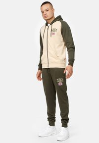 Béžová kapucňová mikina na zips tmavozelenej farby s klokanou kapsou, spárovaná so zodpovedajúcimi tmavozelenými teplákmi s vyšívaným logom.