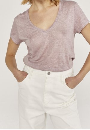 Femme portant un T-shirt violet clair à col en V, rentré dans un pantalon blanc taille haute, les mains dans les poches, sur un fond uni.