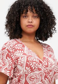 Short-sleeved, V-neck blouse in coral with cream floral pattern. Lightweight fabric, relaxed fit, and subtle textured design details.