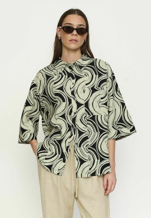 Žena oblečená v nadrozmernej čierno-bežovej vzorovanej košeli na gombíky, béžových nohaviciach, mačacích slnečných okuliaroch a kruhových náušniciach na jednoduchej pozadí.