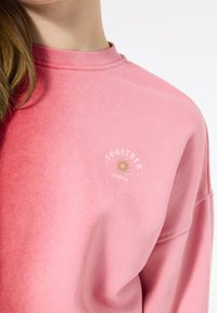 Felpa rosa con scollatura tonda, realizzata in tessuto morbido. Presenta un piccolo logo con il testo "INSIEME" e un'illustrazione di un sole.