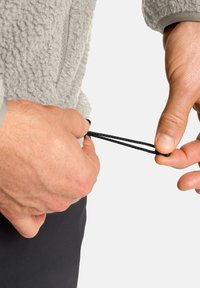 Giacca in fleece grigia con finitura testurizzata. Le mani regolano il cordino nero per una vestibilità perfetta. Primo piano che mostra i dettagli del tessuto e dell'hardware.