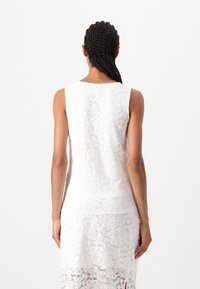 Weißes Spitzenkleid mit ärmellosem Design, das ein Blumenmuster und strukturierte Stoffe aufweist, bis zur Mitte des Oberschenkels reicht und eine figurbetonte Silhouette hat.