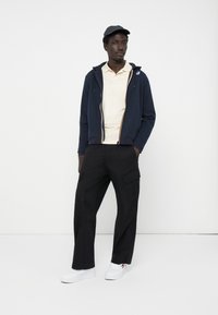 Marineblå zip-up hoodie med hvide og gule detaljer, lys beige polo, sorte cargo-bukser og hvide sneakers, sat mod en ensfarvet baggrund.