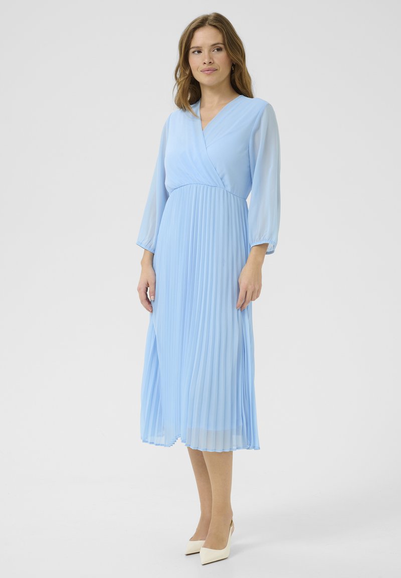 Svijetloplava midi haljina s naborima, dizajnom omote, dugim rukavima i glatkom teksturom, uparen a sa kremasto bijelim potpeticama.