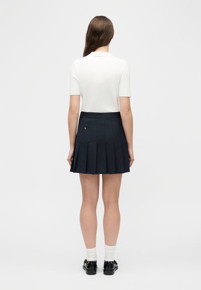 Γυναίκα που στέκεται με την πλάτη, φοράει λευκή μπλούζα με κοντά μανίκια, μπλε πλισέ φούστα με τσέπη, λευκές κάλτσες και μαύρα μοκασίνια.