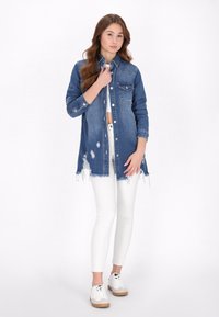 Denim overhemdjasje, blauw, met slijtage-effect, borstzakken en een gebogen zoom. Draagt over witte broek en gecombineerd met witte sneakers.