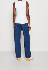 Woman wearing white sleeveless top, blue wide-leg jeans, and cream wooden-soled clogs, standing against a plain light background.