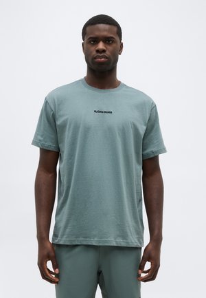 Férfi világoszöld rövid ujjú pólóban, amelyen a mellkas közepén a "BJÖRN BORG" felirat olvasható, egyszínű világos háttér előtt áll.