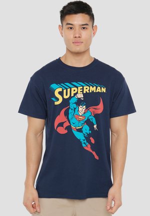 Young man wearing a navy t-shirt with a flying Superman graphic and yellow "SUPERMAN" text on a plain background.