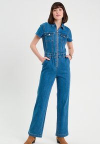Tuta in denim blu con maniche corte, zip frontale, due tasche sul petto e gambe ampie. Texture liscia con leggero elastico.