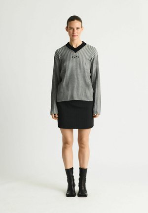 Naine seisab halli soonikkoega kampsuni, musta kraega, musta seeliku ja mustade pahkluu saabastega tavalise valge tausta ees.