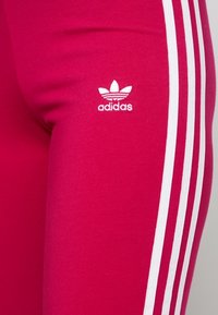 Adidas rosa leggings med vit tre-stripesdesign längs sidorna och en liten vit logotyp på vänster ben. Slät, stretchig material.