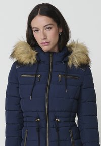 Giacca puffer blu navy con cappuccio foderato in pelliccia, tasche con zip, dettagli in pelle nera e cordini regolabili in vita.