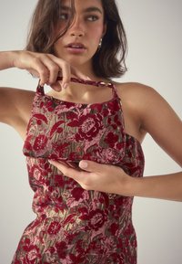 Frau hält eine kleine Handtasche mit Blumenmuster, die zu ihrem trägerlosen Kleid in Rot-, Rosa- und Goldtönen passt.