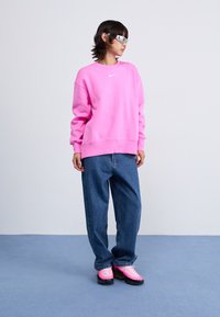 Sweatshirt rose avec un col rond et logo blanc, associé à un jean en denim bleu ample et des chaussures de sport roses avec des accents noirs.