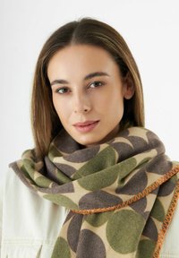Jeune femme aux cheveux bruns raides portant une veste beige et une grande écharpe avec des motifs circulaires verts et bruns, surpiqûres orange.