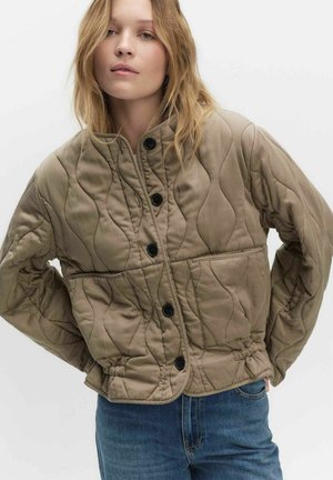 Jonge vrouw met licht haar, gekleed in een beige doorgestikt jasje met zwarte knopen en een blauwe spijkerbroek, tegen een effen achtergrond.