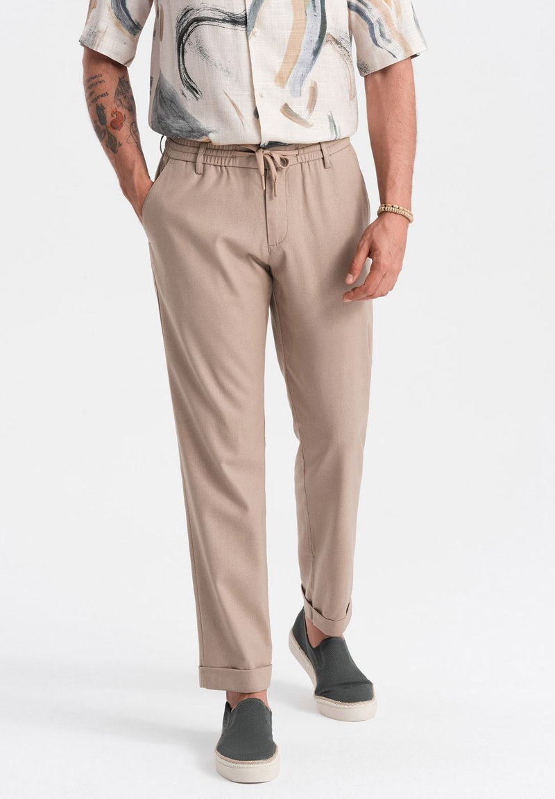 Beige, lässige Hose mit Kordelzugbund, umgeschlagenem Saum und leichtem Stoff. Kombiniert mit grauen Slippern.