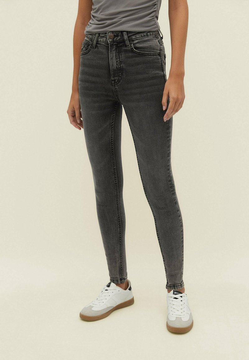 Jeans skinny negros con una cintura alta, que cuentan con cinco bolsillos y un ligero desvanecido. Combinados con zapatillas blancas y una camiseta gris ajustada.