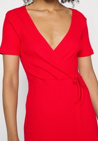 Czerwona, żebrowana sukienka z głębokim dekoltem w kształcie litery V, krótkimi rękawami oraz fasonem kopertowym. Posiada wiązanie w talii umożliwiające regulację dopasowania.