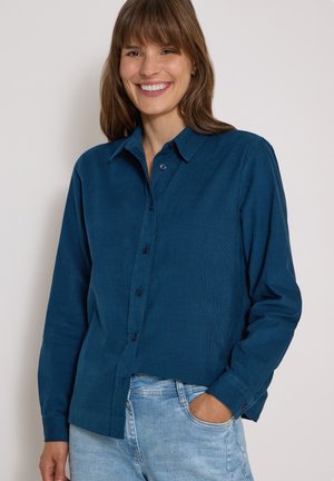 Femme souriante portant une chemise bleu foncé à boutons et un jean bleu clair, une main dans la poche, sur un fond uni.