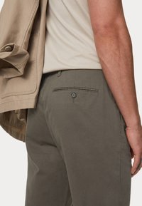 Baumwollhosen in Olivgrün mit einer Gesäßtasche und Knopfverschluss. Eine beigefarbene Jacke lässig über die Schulter gelegt. Einfaches, hellfarbiges Hemd.