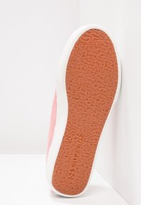 Semelle d'une chaussure de sport rose avec une semelle en caoutchouc brun texturé et une bordure blanche, positionnée verticalement contre un mur d'angle blanc.