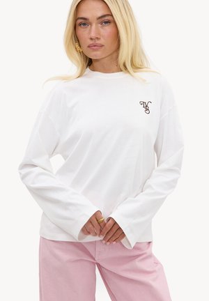 Femme aux longs cheveux blonds porte une chemise blanche à manches longues avec un petit logo sur la poitrine et un pantalon rose clair, debout devant un fond blanc.