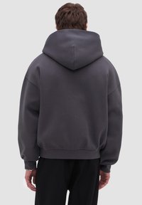 Persona vista desde atrás con una sudadera con capucha de gran tamaño de color gris oscuro y pantalones negros sobre un fondo liso de color gris claro.