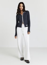 Cardigan zippé bleu marine à bordure blanche, associé à un pantalon blanc à jambes larges. Tissu en maille texturé, design de style sportif, et chaussures noires à motif.