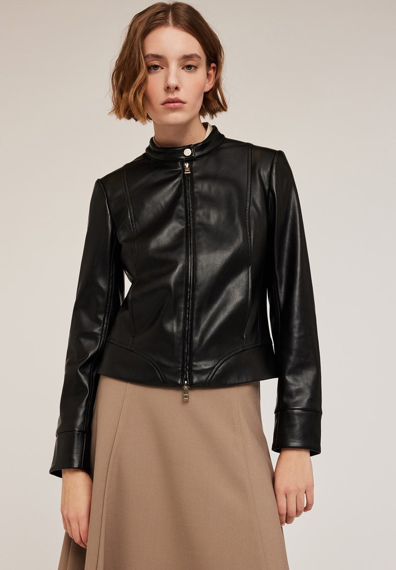 Giubbotto di pelle nera con colletto stand-up, chiusura con zip, design aderente e dettagli di cucitura sottili, abbinato a una gonna beige.