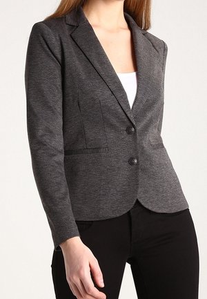 Vrouw draagt een getailleerde donkergrijze blazer met twee knopen over een wit topje en zwarte broek, staand tegen een effen achtergrond.