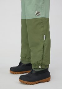 Grüne und hellgrüne wasserdichte Hosen mit elastischen Bündchen, seitlichem Logo und Druckknopfverschluss. Kombiniert mit schwarzen Gummistiefeln und braunen Sohlen.