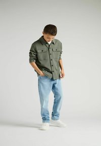 Camicia verde con bottoni, dotata di due tasche frontali, abbinata a jeans azzurri chiari e sneakers bianche. Tessuto morbido e vestibilità comoda.