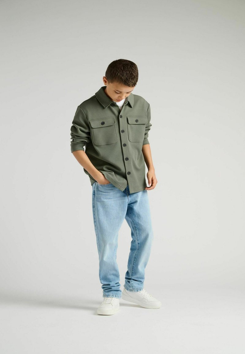 Camicia verde con bottoni, dotata di due tasche frontali, abbinata a jeans azzurri chiari e sneakers bianche. Tessuto morbido e vestibilità comoda.