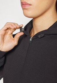 Haut à zip noir avec un tissu lisse et texturé. Dispose d'une capuche ajustée et de deux tirettes zippées au niveau du cou pour un réglage.