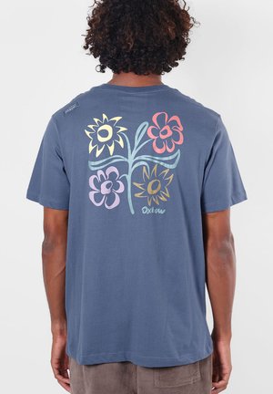 Personne aux cheveux bouclés portant un t-shirt bleu avec quatre grandes illustrations colorées de fleurs dans le dos et le mot "Oxtow."
