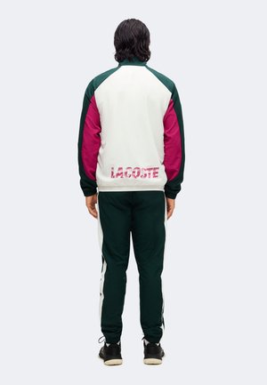 Hombre con cabello oscuro de espaldas, vistiendo un chándal Lacoste blanco, verde y rosa con zapatos negros sobre un fondo blanco.