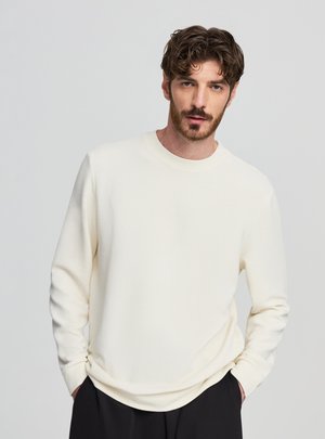 Uomo con capelli ricci castani e barba, indossa una camicia a maniche lunghe color avorio con trama e pantaloni neri, mani in tasca, sfondo neutro.