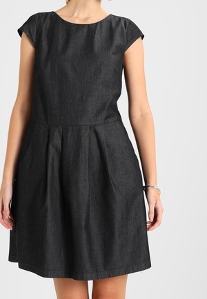 Kobieta w krótkiej sukience o ciemnoszarym kolorze, sięgającej do kolan, z plisami w talii, stojąca na tle jednolitego, jasnego tła.