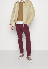 Chaqueta beige con cremallera, suéter de punto marrón, camisa blanca debajo, pantalones burdeos y zapatillas altas marrones con suelas blancas.