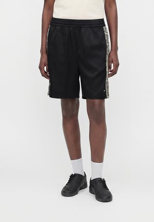 Person iført sorte knælange shorts med mørke camouflagemønstrede sidepaneler, hvide sokker og sorte sneakers, stående mod en ensfarvet lys baggrund.