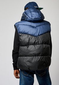 Chaleco hinchado azul y negro con capucha, que presenta una textura acolchada y detalles de cremallera. Llevado sobre una camiseta de manga larga negra.