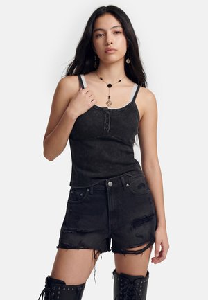 Ung kvinde med langt mørkt hår bærer en sort ærmeløs top, slidte sorte shorts, snørebånds sorte støvler og guldsmykker.