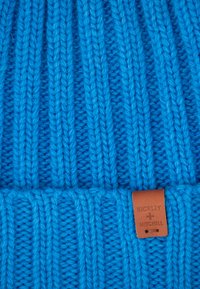 Niebieska czapka dzianinowa z prążkowanym wzorem, z skórzaną metką oznaczoną "BICKLEY + MITCHELL" z boku. Miękki, gruby materiał włóczkowy.