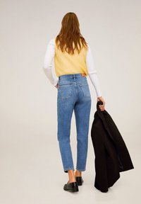 Korkeavyötäröiset siniset farkut suoran lahkeen kanssa, vaaleaa denim-kangasta, takataskut, yhdistettynä keltaiseen neuleeseen ja mustaa bleiseriä pidellen.