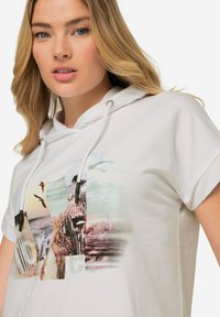 Sudadera blanca con cordón, que presenta una impresión de collage fotográfico en colores suaves. Corte relajado, mangas cortas y textura de algodón suave.