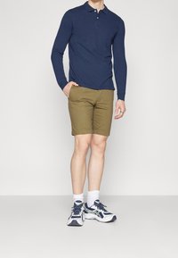 Námořnická polokošile s dlouhým rukávem, khaki šortky a bílé sportovní tenisky s modrými akcenty. Model stojí s jednou rukou v kapse.